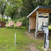 Rastplatz Hopfenberg  Rastplatz mit überdachten Infotafeln, zwei Liegebänken und einer Ladesäule, umgeben von Bäumen.