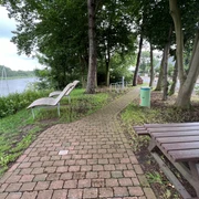 Rastplatz Heimsen  Ein schmaler Pflasterweg führt zu Holzliegen und einem Tisch inmitten von Bäumen am Flussufer.