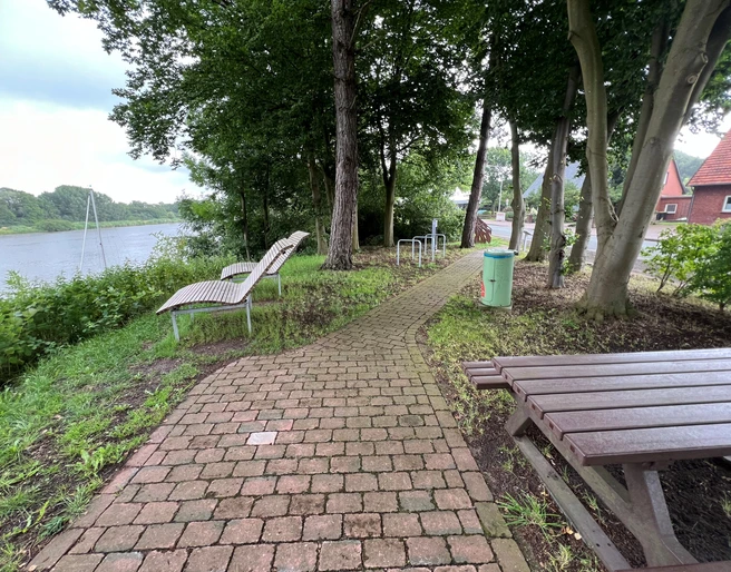 Rastplatz Heimsen  Ein schmaler Pflasterweg führt zu Holzliegen und einem Tisch inmitten von Bäumen am Flussufer.