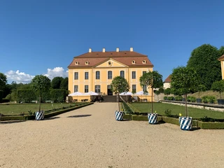 Blick zum Schloss  Gelbes Barockgebäude des Barockgarten Großsedlitz mit rotem Dach, umgeben von gepflegtem Garten und Bäumen in blau-weißen Töpfen, unter klarem blauem Himmel.