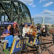 Rikolonia Fünf stilvoll gekleidete Personen sitzen in modernen Rikschas vor der Hohenzollernbrücke in Köln. Die Sonne scheint, der Himmel ist blau mit weißen Wolken. Five stylishly dressed people sit in modern rickshaws in front of the Hohenzollern Bridge in Cologne. The sun is shining, the sky is blue with white clouds.