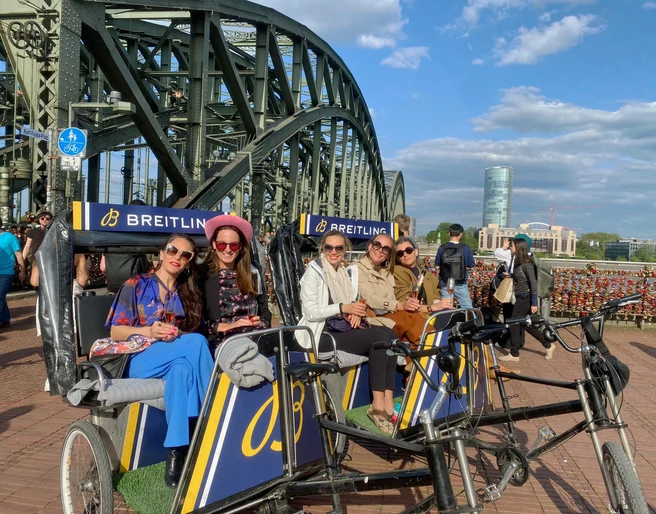 Rikolonia Fünf stilvoll gekleidete Personen sitzen in modernen Rikschas vor der Hohenzollernbrücke in Köln. Die Sonne scheint, der Himmel ist blau mit weißen Wolken. Five stylishly dressed people sit in modern rickshaws in front of the Hohenzollern Bridge in Cologne. The sun is shining, the sky is blue with white clouds.