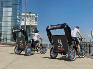 Rikolonia Zwei Rikscha-Fahrer fahren nebeneinander auf einer gepflasterten Promenade am Rhein entlang.Two rickshaw drivers ride side by side on a cobbled promenade along the Rhine.