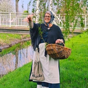 Auguste  Eine Frau im historischen Kostüm, mit Kopftuch und Schürze, hält einen Korb. Sie steht lachend auf einer grünen Wiese neben dem Augustfehn-Kanal und einem Baum, im Hintergrund ist eine weiße Brücke zu sehen.