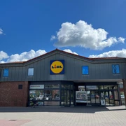 Lidl.jpg Ein Lidl Supermarkt mit grauem Dach und blauem Logo vor einem klaren Himmel mit Wolken.