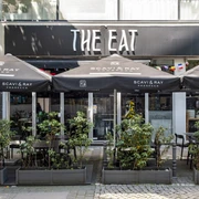The Eat Das Bild zeigt ein Restaurant namens "The Eat" mit mehreren Tischen unter schwarzen Sonnenschirmen. Der Eingangsbereich ist mit Pflanzen dekoriert.The image shows a restaurant named "The Eat," featuring several tables underneath black sun umbrellas. The entrance area is adorned with plants.