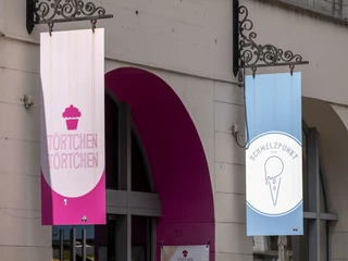 Toertchen Toertchen Zwei Schilder hängen über dem Eingang eines Gebäudes. Links ein Schild mit der Aufschrift "Törtchen Törtchen" auf rosa Hintergrund, rechts ein Schild für "Schmelzpunkt" mit einem Eis-Symbol auf blauem Hintergrund. Two signs hang above the entrance of a building. On the left is a sign reading "Törtchen Törtchen" against a pink background, and on the right, a sign for "Schmelzpunkt," featuring an ice cream symbol on a blue background.