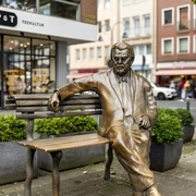 Willy Millowitsch memorial Das Bild zeigt eine bronzene Statue von Willy Millowitsch, die auf einer Parkbank sitzend dargestellt ist. Die Statue befindet sich in einer urbanen Umgebung, flankiert von Blumenbeeten und Gebäuden.The picture shows a bronze statue of Willy Millowitsch sitting on a park bench. The statue is located in an urban setting, flanked by flowerbeds and buildings.