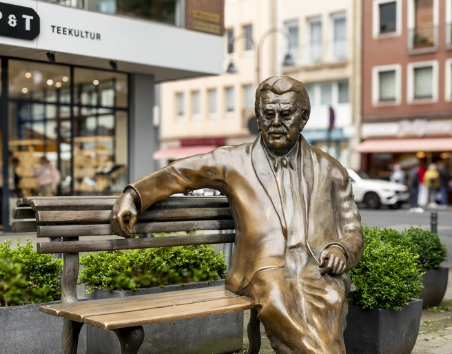 Willy Millowitsch memorial Das Bild zeigt eine bronzene Statue von Willy Millowitsch, die auf einer Parkbank sitzend dargestellt ist. Die Statue befindet sich in einer urbanen Umgebung, flankiert von Blumenbeeten und Gebäuden.The picture shows a bronze statue of Willy Millowitsch sitting on a park bench. The statue is located in an urban setting, flanked by flowerbeds and buildings.