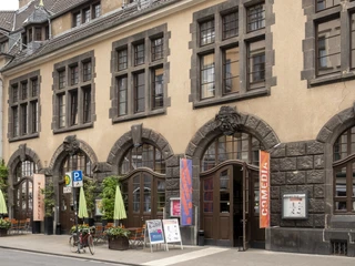 Comedia Das Bild zeigt die Fassade des historischen Gebäudes der Comedia in Köln, ein kulturelles Zentrum mit großen Sprossenfenstern und dekorativen Steinornamenten. Vor der Gebäudezeile laden Schirme und Sitzmöbel unter freiem Himmel zum Verweilen ein.The picture shows the façade of the historic Comedia building in Cologne, a cultural center with large mullioned windows and decorative stone ornaments. In front of the row of buildings, umbrellas and outdoor seating invite you to linger.