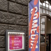 Comedia  Der Eingang der Comedia-Theater in Köln zeigt bunte Fahnen und ein beleuchtetes Plakat, das vorbeigehende Menschen dazu einlädt, die lebendige Kulturszene zu entdecken.The entrance to the Comedia theaters in Cologne displays colorful flags and an illuminated poster inviting passers-by to discover the lively cultural scene.