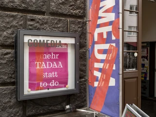 Comedia  Der Eingang der Comedia-Theater in Köln zeigt bunte Fahnen und ein beleuchtetes Plakat, das vorbeigehende Menschen dazu einlädt, die lebendige Kulturszene zu entdecken.The entrance to the Comedia theaters in Cologne displays colorful flags and an illuminated poster inviting passers-by to discover the lively cultural scene.