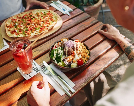 Stadtgarten Ein bunt gedeckter Holztisch zeigt einen sommerlichen Tisch im Freien mit einem frischen Salat und Pizza.A colorfully set wooden table showcases a summery outdoor dining scene, featuring a fresh salad and pizza.