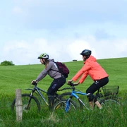 Raderlebnis Schieder-Schwalenberg Zwei Radfahrer mit Helmen genießen eine Fahrt auf einem grünen Weg bei bewölktem Himmel.