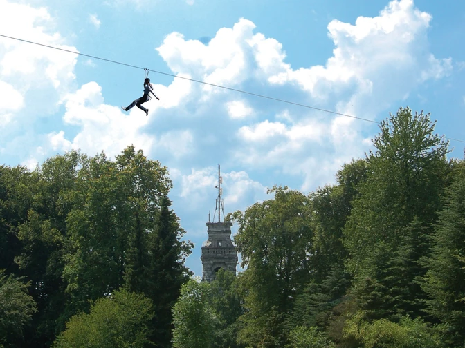 Seilbahn mit Bismarckturm im Waldkletterpark Velbert-Langenberg.jpg Eine Person schwebt mit einer Seilbahn über Baumwipfel vor dem Bismarckturm im Kletterpark.