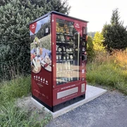 Proviantomat Standort in Pötzscha/Stadt Wehlen Ein roter Verkaufsautomat steht auf einem Betonfundament in einer grünen, ländlichen Umgebung, gefüllt mit verschiedenen Lebensmitteln.