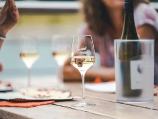 Wine festival on the Rhine Das Bild zeigt eine stilvolle Weinszene auf einem Holztisch. Ein Glas Weißwein befindet sich im Fokus, auf dem die Schrift "Weinfest am Rhein" erkennbar ist. Im Hintergrund sind weitere Gläser und unscharfe Umrisse von Personen zu sehen, die eine entspannte und gesellige Atmosphäre vermitteln. Der Tisch ist mit Tellern und einer eingekühlten Flasche bedeckt, was die gesellige Stimmung dieses Weinfestes unterstreicht.The picture shows a stylish wine scene on a wooden table. A glass of white wine is in focus, on which the words "Weinfest am Rhein" can be seen. More glasses and blurred outlines of people can be seen in the background, conveying a relaxed and convivial atmosphere. The table is covered with plates and a chilled bottle, emphasizing the convivial atmosphere of this wine festival.