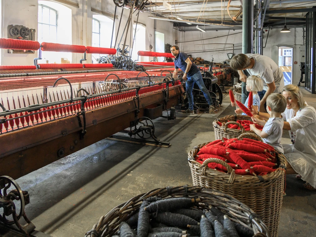 bramsche-tuchmacher-museum(c)tol-foto-jurjen-drenth (11).JPG Historische Weberei mit Besuchern, roten Garnspulen, alten Maschinen in einem lichtdurchfluteten Raum.