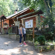 Ibbenbüren-Almhütte-1600.jpg Zwei Frauen stehen vor einer Informationstafel vor einer Holzhütte im Wald an einem sonnigen Tag.