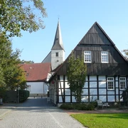 Lienen_Haus des Gastes_Foto_Gemeinde Lienen.JPG Fachwerkhaus mit weißer Fassade und dunklen Holzbalken, im Hintergrund eine Kirche mit hoher Turmspitze.