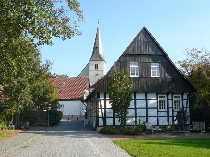 Lienen_Haus des Gastes_Foto_Gemeinde Lienen.JPG Fachwerkhaus mit weißer Fassade und dunklen Holzbalken, im Hintergrund eine Kirche mit hoher Turmspitze.
