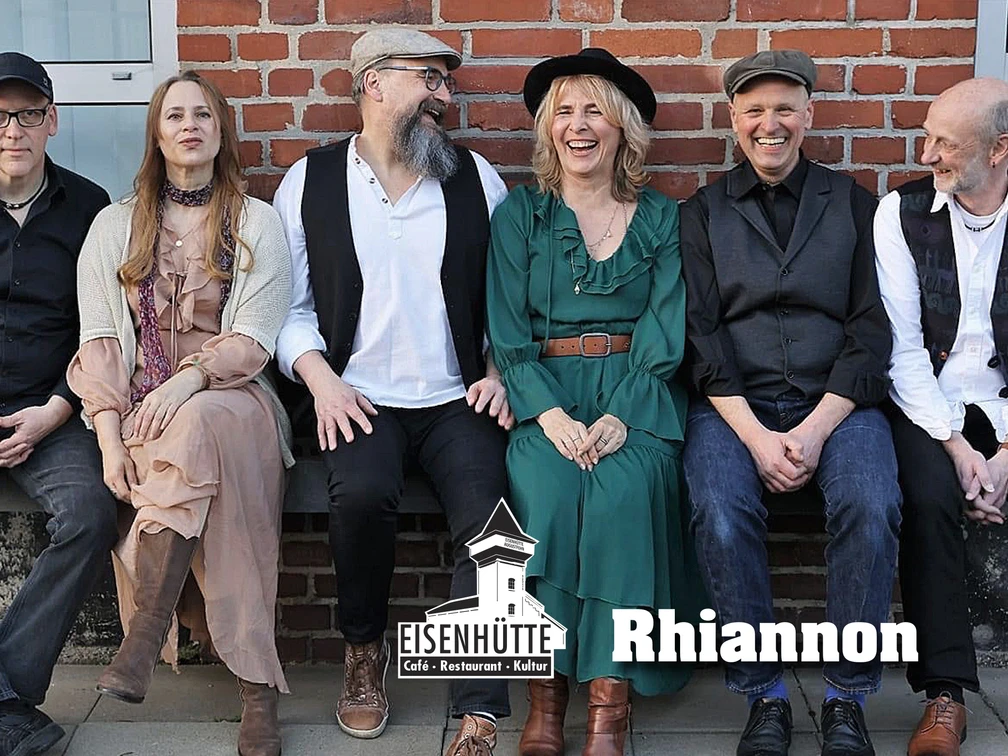 6 Rhiannon.jpg Sechs Mitglieder der Fleetwood‑Mac‑Coverband Rhiannon sitzen entspannt vor einer Backsteinwand auf einer niedrigen Mauer, lachend oder lässig in die Kamera – darunter zwei Frauen und vier Männer – mit dem Schriftzug ‚Eisenhütte – Café · Restaurant · Kultur‘ und dem Bandnamen ‚Rhiannon‘.