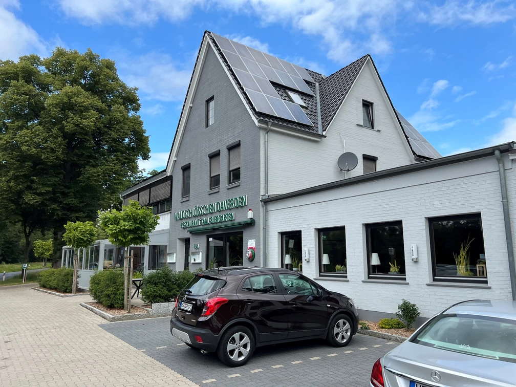 Waldschlößchen Daverden.jpg Ein zweistöckiges Gebäude mit weißen Wänden, grauem Dach und Solarpanelen, im Vordergrund geparkte Autos.
