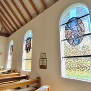 Hövelsenner Kapelle Drei bunte Glasfenster werfen farbenfrohe Lichtreflexe in die Holzkirche mit Holzdeckenbalken.
