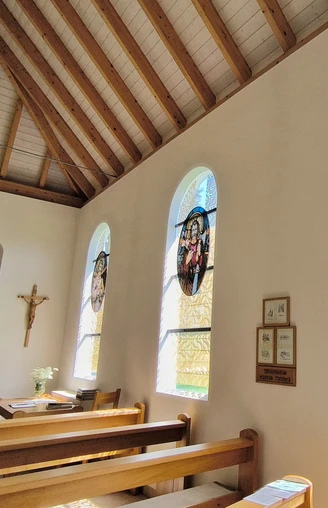 Hövelsenner Kapelle Drei bunte Glasfenster werfen farbenfrohe Lichtreflexe in die Holzkirche mit Holzdeckenbalken.