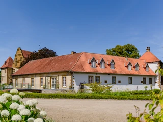 _BBB7003.jpeg Historisches Gut mit roten Ziegeldächern, umgeben von Grünflächen und blauem Himmel im Hintergrund.