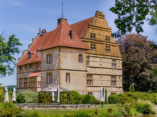 _BBB7026.jpeg Historisches Schlossgebäude mit markantem roten Dach, umgeben von Garten und Sonnenschirmen.