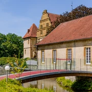 _BBB7013.jpeg Schloss mit rotem Ziegeldach, umgeben von Bäumen und Wassergraben, bei sonnigem Wetter.