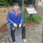 20231106_102208.jpg Statue eines Lokführers in traditioneller Uniform, sitzend auf Bank in belebter Fußgängerzone.