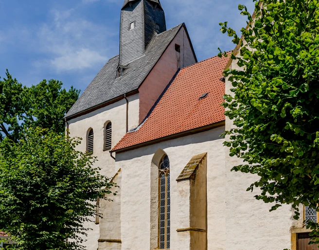 Eine historische Kirche mit spitzem Turm und rotem Ziegeldach, umgeben von grünen Bäumen.