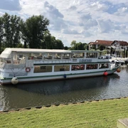 MS Stadt Verden - Flotte Mittelweser Ein weißes Passagierschiff auf einem ruhigen Fluss neben einem kleinen Hafen mit Gebäuden im Hintergrund.