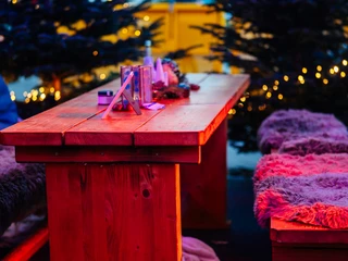 Eine gemütliche Sitzbank in der Gänseliesels Weihnachtslounge Rustikaler Holztisch mit Fell überzogenen Bänken, stimmungsvoll beleuchtet, umgeben von Tannen.