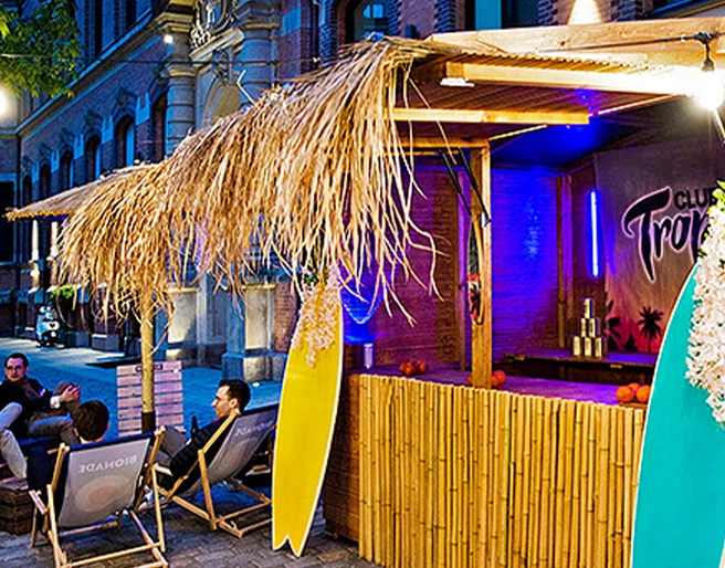 mSa Club Tropicana Sommerfest Sommerfeeling im Club Tropicana: Holzunterstand mit Surfbrettern, im Hintergrund sitzen Personen im Kreis auf Liegestühlen mit Getränken in der HandSummer feeling at Club Tropicana: wooden shelter with surfboards, people sitting in a circle on sun loungers with drinks in their hands in the background