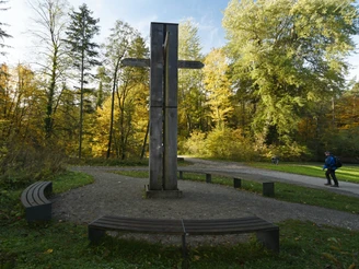 Holzkreuz auf einer kreisförmigen Plattform; umgeben von Bänken und Herbstlaub im Wald.