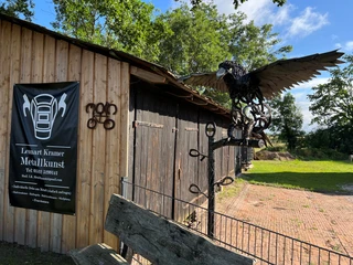 Metallskulptur eines Adlers, breitet seine Flügel aus, vor einer rustikalen Holzscheune platziert.