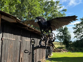Skulptur Adler Eiserne Adler-Skulptur auf einem Ast vor einer Holzhütte, mit blauen Himmel und Bäumen im Hintergrund.