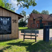 Skulptur Adler Metallene Skulptur eines Adlers auf einem Holzpfosten vor einem Gebäude, umgeben von grünem Gelände.