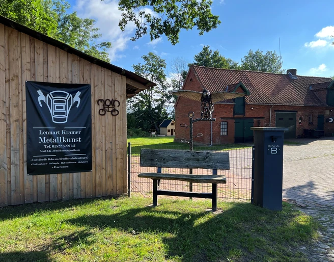 Skulptur Adler Metallene Skulptur eines Adlers auf einem Holzpfosten vor einem Gebäude, umgeben von grünem Gelände.