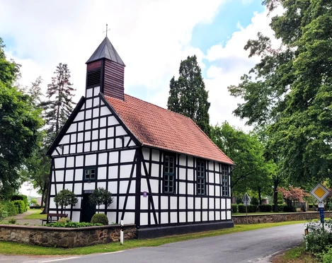 Fachwerkkapelle mit rotem Ziegeldach und kleinem Turm, umgeben von grünen Bäumen an einer Straße.