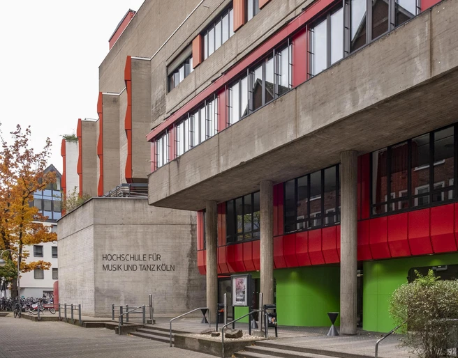 University of Music and Dance Markantes Gebäude der Hochschule für Musik und Tanz Köln mit roten und grünen Akzenten an der Fassade, umgeben von Bäumen und urbaner Architektur.Striking building of the Cologne University of Music and Dance with red and green accents on the façade, surrounded by trees and urban architecture.