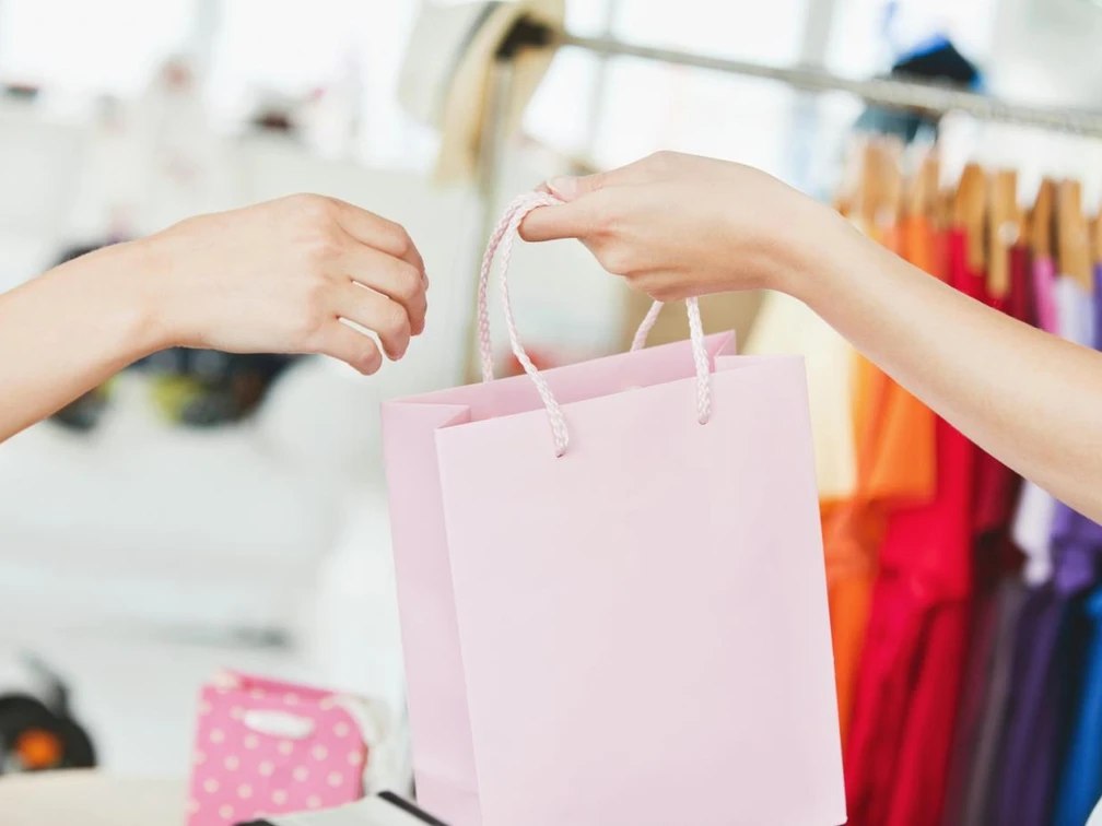 Eine rosa Einkaufstasche wird zwischen zwei Personen vor einer bunten, mit Kleidung gefüllten Kulisse übergeben.