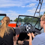 Bootsvermietung NienBoot Zwei Personen fahren ein Motorboot auf einem ruhigen Fluss, umgeben von Bäumen und blauem Himmel.