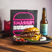 Tisch mit einem Burger auf einem Teller, einer Flasche Bier und einer aufgestellten Karte, auf der "Smash up Burger" steht.