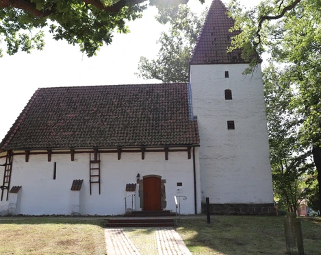 Weiße Kapelle mit markantem Satteldach und seitlichem Turm, umgeben von Bäumen und Natur.