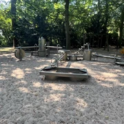 Wasserspielplatz Spielplatz mit Wasserspielen aus Metall, eingerahmt von schattenspendenden Bäumen im Hintergrund.