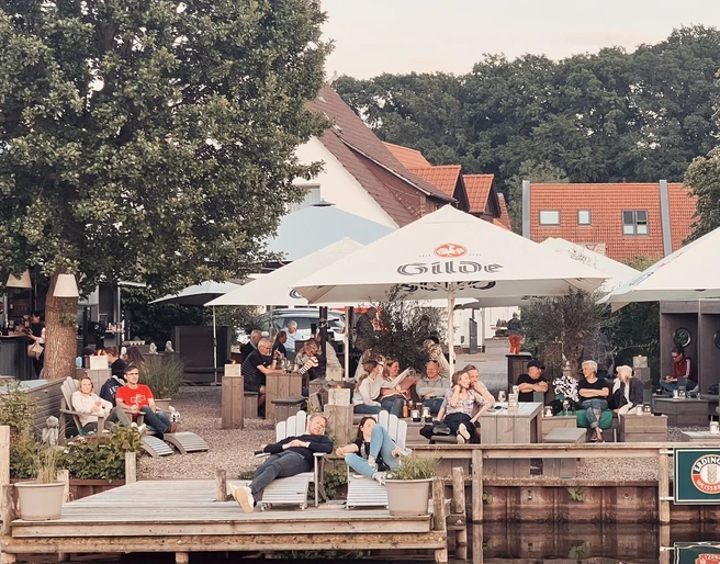 Kranenborgh - Blick auf den Steg Personen entspannen sich im Kranenborgh Biergarten am Wasser unter schattigen Sonnenschirmen.